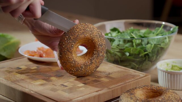 Hand slices seeded bagel with knife on wooden cutting board near greens and ingredients