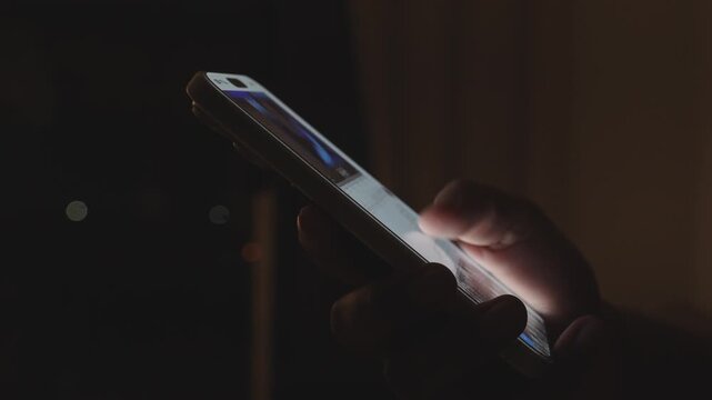 Hand holding smartphone and scrolling social media feed in dark room with desk on background, illuminated by warm tone light. Digital addiction and mindless content consuming