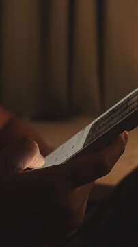 Hand holding smartphone and scrolling social media feed in dark room with desk on background, illuminated by warm tone light. Digital addiction and mindless content consuming