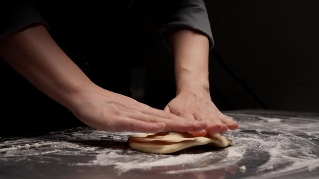 Closeup baker hands pressing and shaping soft dough on floured countertop in dim kitchen, demonstrating artisan homemade bread preparation and rustic cooking technique