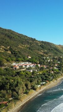 Aerial view of the scenic coastline and hillside with houses in Acciaroli, Italy, under a clear blue sky. Lush greenery and gentle waves create a tranquil Mediterranean atmosphere.