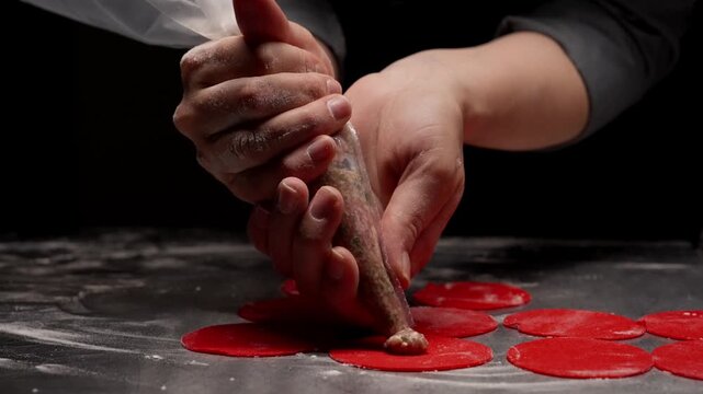 Close-up culinary preparation of red dough circles being carefully filled with seasoned meat mixture using piping technique by skilled hands on dark background