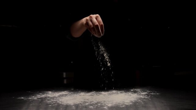 Slow motion hand sprinkling white flour over dark surface in dramatic low key lighting, capturing the delicate motion of baking preparation and culinary artistry