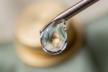 Macro shot of a high-viscosity serum droplet suspended from a precision surgical needle. Subtle micro-bubbles trapped in translucent fluid reflect soft studio lighting against a muted metallic and gol