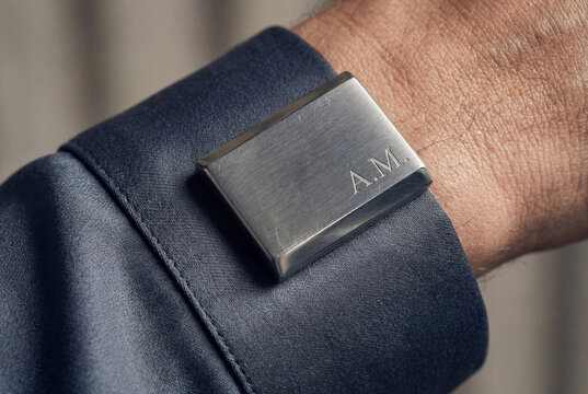 Macro shot of a custom brushed metal cufflink engraved with A.M. on a dark silk-blend shirt. Capturing micro-scratches on the metallic surface and the subtle sheen of high-thread-count textile. Pure a