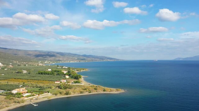 Aerial view of a serene coastline with clear blue water, green fields, and scattered houses near Hermione, Greece under a partly cloudy sky.