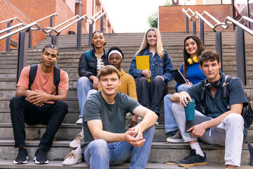 Fototapeta premium Group of diverse university students with backpacks and books sitting on outdoor stairs, showing friendship and education