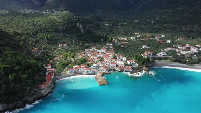 Aerial view of a picturesque coastal village with white houses and red roofs by turquoise waters near Monemvasia, Greece. Lush green hills surround the charming Mediterranean settlement.