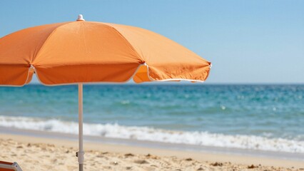 Fototapeta premium An orange beach umbrella provides shade on a sandy shore with the blue ocean and clear sky in soft focus.