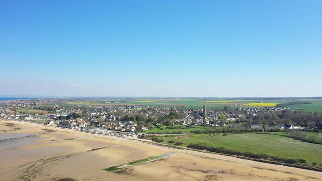 Wide aerial shot of Juno Beach and the coastal village of Bernieres sur Mer, Normandy, with sandy shoreline, residential houses, and green fields under a clear blue sky.