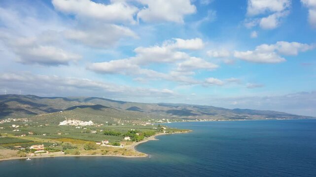 Aerial view of a serene coastline with clear blue water, rolling hills, and scattered houses near Hermione, Greece, under a partly cloudy sky.