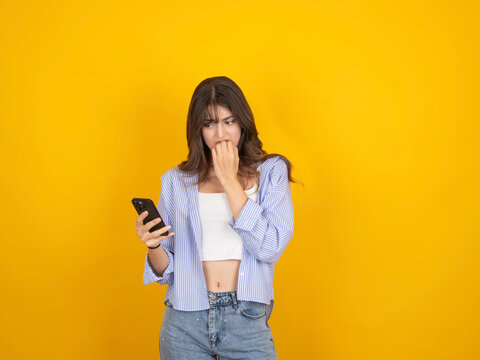 Young woman looking worried while checking smartphone, biting nails in anxiety. Female using mobile phone isolated on yellow background with copy space, concept of stress, bad news, social media or no