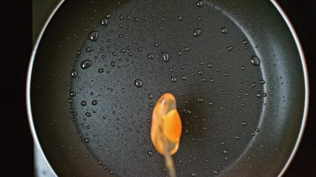 Egg splatters into a sizzling hot pan, white albumen spreading as yolk remains centered, droplets of oil splash around the frying surface