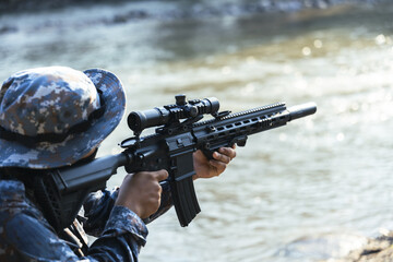 Obraz premium Focused soldier in tactical camouflage uniform aiming modern black assault rifle with scope. special force operative is positioned near flowing river, ready for combat
