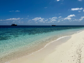 Fototapeta premium Beautiful tropical beach with white sand and turquoise clear ocean water on a sunny day. Idyllic sand beach shoreline with crystal clear sea water and safari boats on the horizon. 