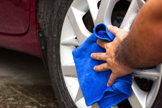 Car Wheel and Alloy Cleaning, A hand uses a bright blue microfiber cloth to carefully wipe and polish the silver alloy rim of a red vehicle.