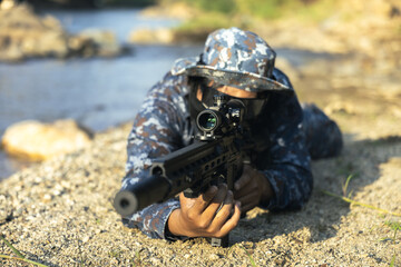 Obraz premium Special force soldier with focused look, in prone position aiming sniper rifle. serviceman in camouflage uniform on covert military operation outdoors by river
