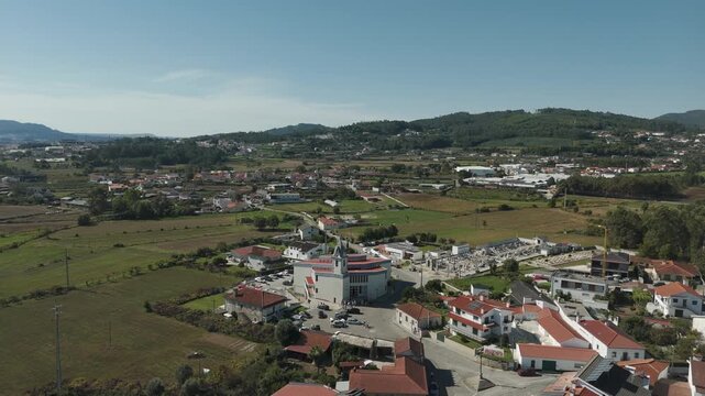 Drone aerial view of Igreja Paroquial de Lij&oacute; in Barcelos Portugal featuring parish church cemetery surrounding farmland village houses and green hills under clear sky