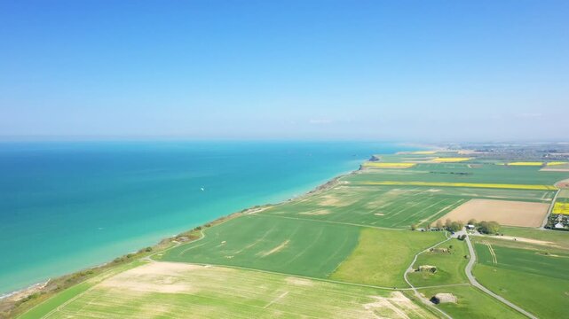 Drone shot of green fields and dramatic coastline at Longues sur Mer, Normandy, France, under a clear blue sky. The turquoise sea contrasts with the patchwork of farmland along the coast.