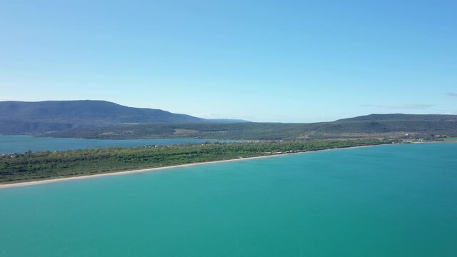 Drone shot of the turquoise sea and lush coastline at Isola Varano Nature Reserve in Italy, with mountains in the background under a clear blue sky.