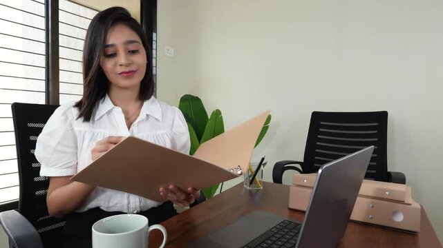 Closeup of happy successful working young Asian business woman boss manager sits on chair at workplace desk hold file analysing documents paper with satisfied face expression.