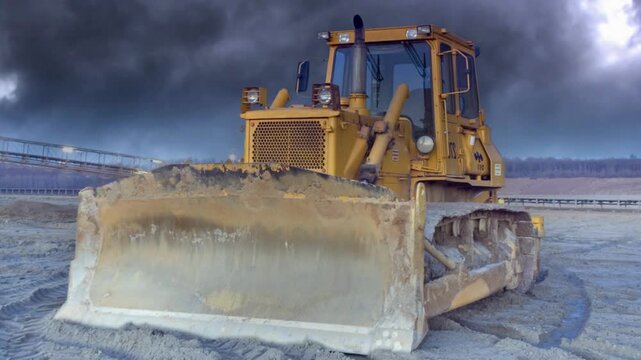 Large yellow bulldozer on a construction site with dirt in front under a dramatic dark and cloudy sky signifying heavy industry