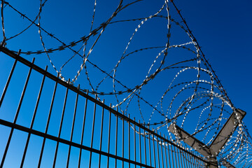 High barrier fence at a security area of D&uuml;sseldorf Airport on a sunny day, isolated against a blue sky. Barbed wire coils with shiny, razor-sharp metal blades above the metal mesh fence. A barrier th