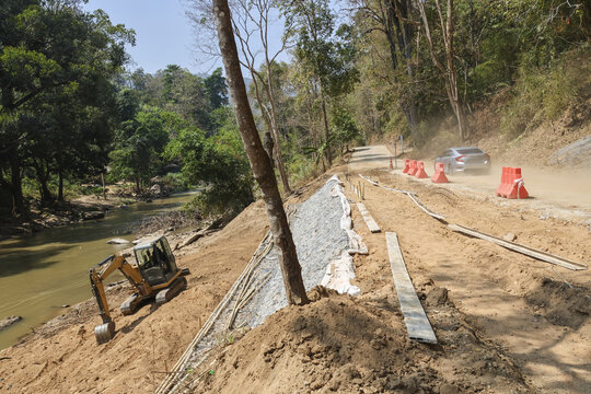Rural road construction with an excavator at river bank. Civil engineering work for slope stabilization and infrastructure development in scenic mountain forest landscape