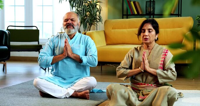 Indian senior couple doing omkar pranayama or chanting mantra or yoga at home in the morning sitting with folded legs in modern living room, practicing mindful breathing for health