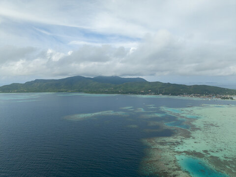 Aerial view of the island's dark blue waters meeting the vibrant turquoise reef, with lush green hills under a cloudy sky, Karimunjawa, Central Java, Indonesia.