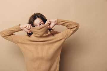 Fototapeta premium Young woman in beige knitted sweater pulls collar over face, looking sideways with playful expression. Neutral background highlights cozy autumn fashion and casual lifestyle concept.