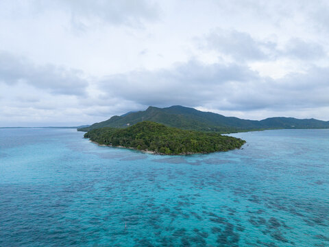 Aerial view of lush green island meeting the turquoise sea under a cloudy sky near Tanjung Gelam Beach, Karimunjawa, Central Java, Indonesia.