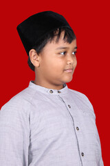 Young Indonesian boy wearing a traditional black peci cap and a grey Islamic koko shirt, looking thoughtfully to the side against a vibrant red background.