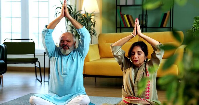 Indian senior couple doing omkar pranayama or yoga at home in the morning sitting with folded legs in modern living room chanting aum, practicing mindful breathing exercise for health