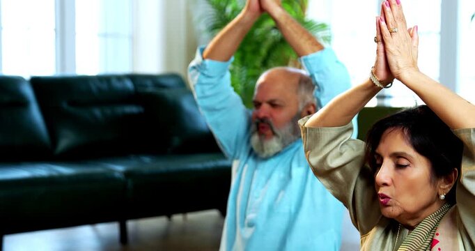 Indian senior couple doing omkar pranayama or yoga at home in the morning sitting with folded legs in modern living room chanting aum, practicing mindful breathing exercise for health