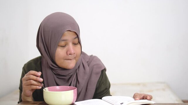 Mature Asian woman in hijab eating instant noodles from a bowl alone in her bedroom. Muslim lady enjoying hot spicy ramen at home