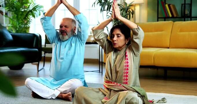 Indian senior couple doing omkar pranayama or yoga at home in the morning sitting with folded legs in modern living room chanting aum, practicing mindful breathing exercise for health