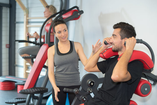 young friendly female coach assisting man using machine in gym