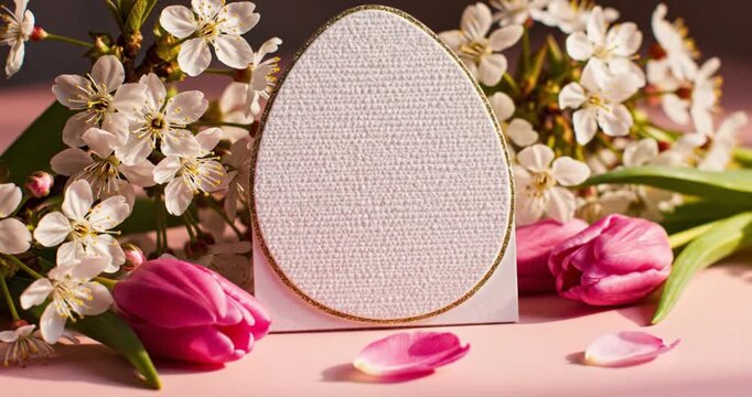Elegant egg-shaped card, golden border, textured white surface. Framed by white blossoms and pink tulips. Soft light, pink background, festive spring mood. Ideal for greetings, celebrations, copy spac
