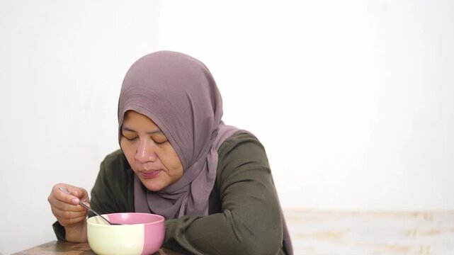 Mature Asian woman in hijab eating instant noodles from a bowl alone in her bedroom. Muslim lady enjoying hot spicy ramen at home