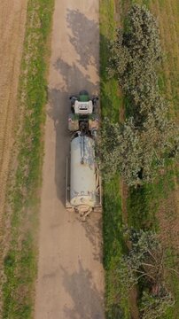 A tractor navigates a dusty road in a serene agricultural landscape, towing a large tank, with rows of crops extending towards the horizon and a tree lining.