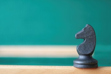 A black wooden knight chess piece stands on a wooden edge against a blurred green background. Concept of non-standard strategy. © Helga P-A.
