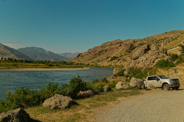 Vehicle By River. Lone Road Traveler Unloads Gear Beside Mountain Stream Under Bright Midday Sun © Val MAK