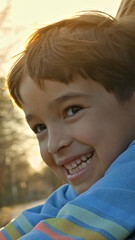 Obraz premium Close-up shot of a young boy smiling brightly in a blue striped sweater, with warm sunlight enhancing the background's serene outdoor setting, suggesting a joyful, carefree lifestyle.