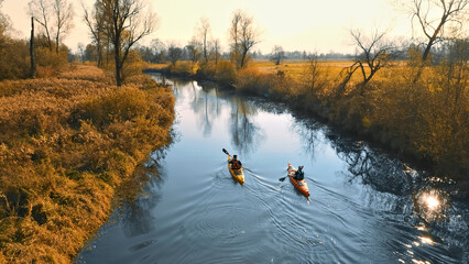 Two kayakers navigate a serene, sunlit river surrounded by autumn foliage. The calm water reflects...