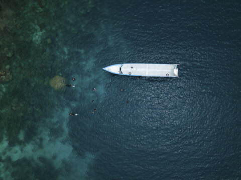 Aerial view of a pristine white boat surrounded by swimmers in the deep blue sea near the Cemara Kecil Snorkeling Spot, Karimunjawa, Central Java, Indonesia.