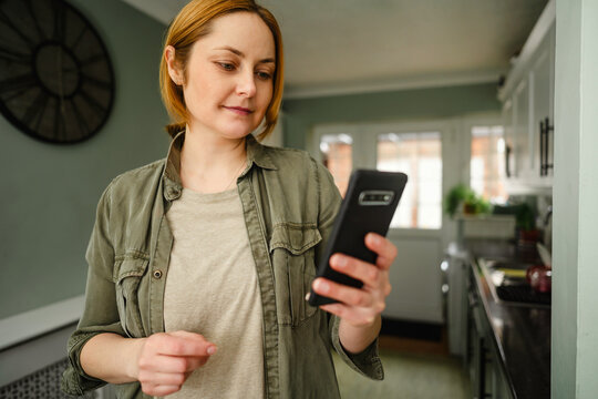 Woman using smartphone in kitchen for online browsing and communication