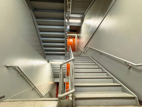 Stairs inside a building lead to an upper level while another set descends to a lower area, with walls painted in light colors and bright lights overhead