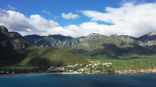 Aerial view of a coastal village nestled between lush green hills and dramatic mountains near Monemvasia, Greece, with turquoise waters in the foreground and a partly cloudy sky.