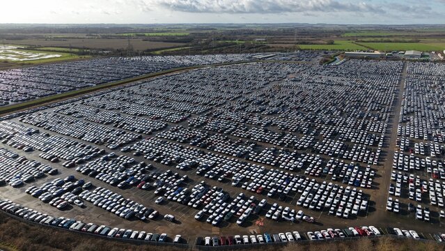Aerial view of a vast expanse of tightly packed cars glistens under an overcast sky, creating a sea of monochrome against the backdrop of distant fields, Grimsby, United Kingdom.